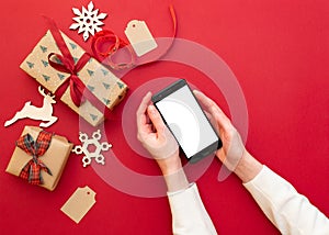 women`s hands holding phone on red background with Christmas decorations and gifts. Xmas and Happy New Year composition. Flat lay