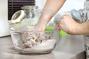 Women`s hands make dough for baked goods. The glass bowl with ingredients stands on the kitchen counter.