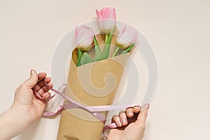 Women`s hands pack a bouquet of fresh tulips using brown eco-paper and satin ribbon, flatlay composition on a beige background