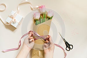 Women`s hands pack a bouquet of fresh tulips using brown eco-paper and satin ribbon, flatlay composition on a beige background