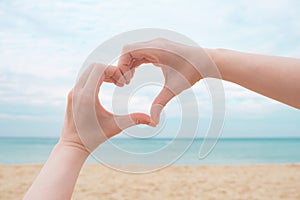 Women's hands in the shape of a heart on a background of sand, blue sea and sky