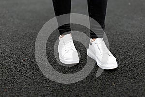 Women`s legs in black jeans in fashionable white leather sneakers stand on the pavement. Young woman walks down the street