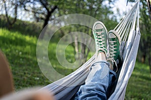 Women's legs shod in green sneakers on a hammock in a summer garden. Summer holidays vacation relaxing in hammock