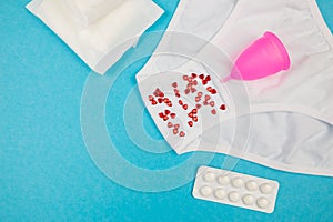 Women`s white panties with red heart confetti, menstrual cup, pills and pads on a blue background. copy space