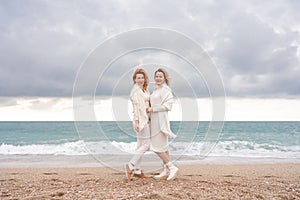 Women sea walk friendship spring. Two girlfriends, redhead and blonde, middle-aged walk along the sandy beach of the sea