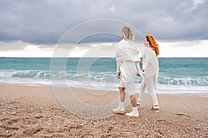 Women sea walk friendship spring. Two girlfriends, redhead and blonde, middle-aged walk along the sandy beach of the sea