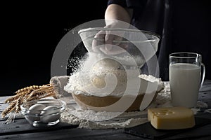 Women sifting flour in bowl on wooden table. Dark background. Dough ingredients.