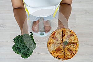 Women stand weighing on the scales. A healthy woman hand holds between a Broccoli and pizza. Deciding to eat foods high in fiber a