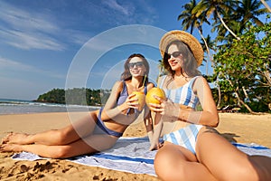 women in striped swimsuits enjoy coconut drinks on sandy beach. Sunbathers relax under tropical sun, clear blue sky