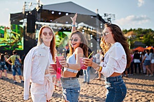 Women toast with cocktails at sunny beachside music event, enjoying performance, friendship, and leisure. Smiling