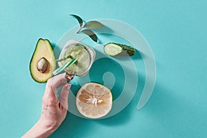 Womens fingers holding ta glass with green smoothie from green vegetables and fruits on a green background with copy