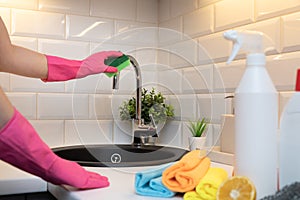 Womens hands cleaning kitchen tap on a black sink with rubber gloves and in front blurred cleaning products for disinfection