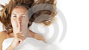 Womens secrets concept. Portrait of a woman lying in bed covered with a blanket holding finger on lips, asking to be quiet,
