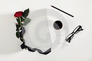 Womens working space with cup of coffee, pencil, empty notebook, glasses, black ribbon and rose flower on white table top view.