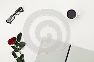 Womens working space with cup of coffee, pencil, empty notebook, glasses and rose flower on white table from above. Flat lay.