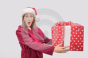 Wondered and amazed young woman in red coat and Christmas hat holding big box of present. Model looks on camera