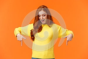 Wondered and excited, amused cute redhead woman in yellow sweater pointing fingers down, look bottom and smiling, check
