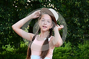 wonderful cheerful young girl with two braids and hat poses against background of green foliage.