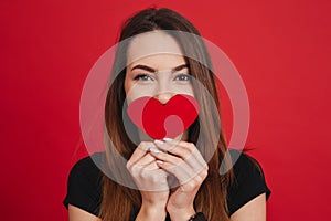 Wonderful happy woman 20s with long brown hair holding paper heart in valentine day at her mouth, isolated over red background