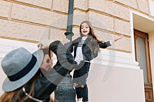 Wonderful little girl enjoying weekend with mother, embracing iron pillar and posing for photo. Young brown-haired lady