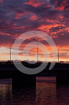 Wonderful sunset sky with colorful clouds and sunbeam over black bridge over river with square frame with reflection of sunlight.