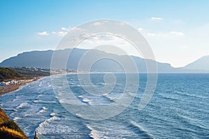 Wonderful view of beach with buildings seashore coastline, sea waves, mountains surrounded by clouds on sunny windy day.