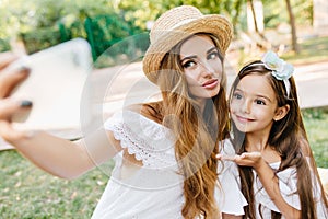 Wonderful young lady with light make-up holding smartphone and making selfie with daughter on nature background