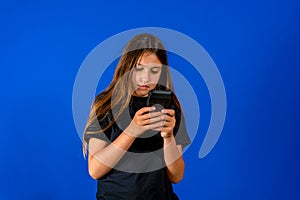 Wondering little cute girl surprised with an interesting information on mobile smartphone. Isolated on blue background.