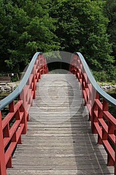 A wood arch bridge, with red side railings, over a lake leading to a forest in Janesville, Wisconsin