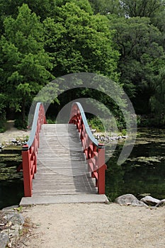 A wood arch bridge, with red side railings, over a lake leading to a forest at the Rotary Botanic Gardens