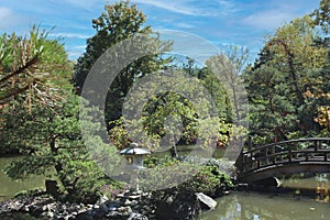 A wood bridge leading to a small island in a lake with evergreens, shrubs and a Japanese stone lantern