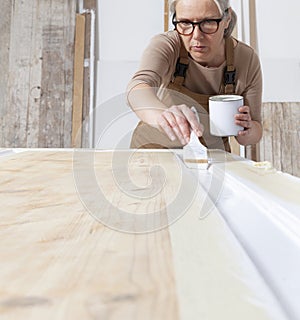 Wood crafts, woman artisan carpenter painting with brush and paint jar white the door in workshop, wearing overall and eyeglasses