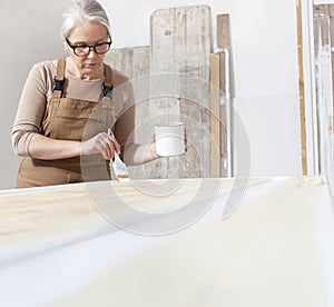 Wood crafts, woman artisan carpenter painting with brush and paint jar white the door in workshop, wearing overall and eyeglasses