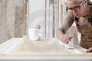 Wood crafts, woman artisan carpenter painting with brush and paint jar white the door in workshop, wearing overall and eyeglasses