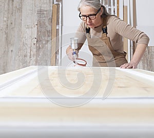 Wood crafts, woman artisan carpenter painting with spray gun paint white the door in her workshop, wearing overall and eyeglasses