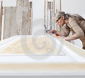 Wood crafts, woman artisan carpenter painting with spray gun paint white the door in her workshop, wearing overall and eyeglasses