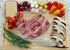 Wood cutting board in kitchen table with beef short ribs, rosemary, mushrooms, tomatoes, cheese, onions and peppers.