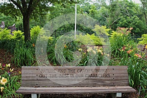 A wood park bench engraved with a quote by Felicity Bryan in front of a garden filled with Daylilies and shrubs