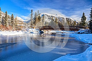 Wood Pedestrian Bridge over Frozen River Ice in Canmore, Canada with Snowcapped Mountain Peaks Landscape blue sky the background