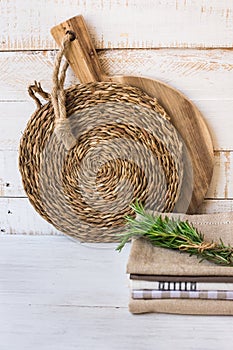 Wood round cutting board, rattan coaster, stack of linen kitchen towels, rosemary twigs on white plank wood