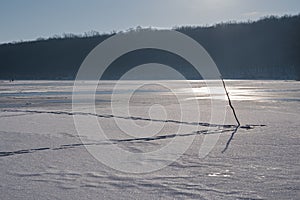 Wood stick frozen in fishing ice hole, fisherman trace path on frozen lake surface after thaw, backlight sun flare winter forest