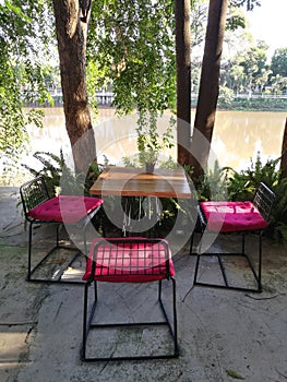 Wood table and chairs with red cushion under tree with sun light in peaceful beautiful garden by river at restaurant backyard, Spr