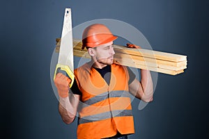 Woodcraft concept. Man, handyman in helmet, hard hat holds handsaw and wooden beams, grey background. Carpenter