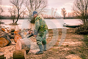 Woodcutter, logger, is cutting firewood, logs of wood, with motor chainsaw near the river