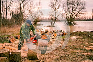 Woodcutter, logger, is cutting firewood, logs of wood, with motor chainsaw near the river