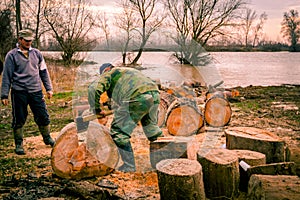 Woodcutter using axe to split large trunk of hardwood, chop firewood at river bank