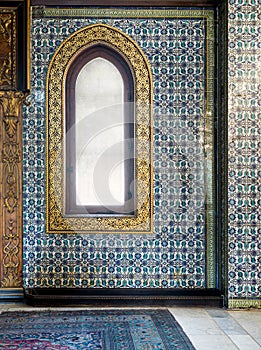 Wooden arched window framed by golden floral pattern ornaments over ceramic tiles wall with floral blue patterns