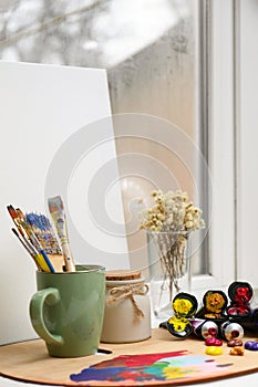 Wooden artist`s palette with colorful paints, brushes and canvas on white windowsill