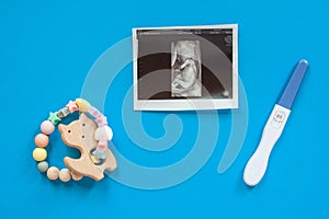 Wooden baby teether with beads, ultrasound image and pregnancy test. Minimalistic flat lay with a wooden toys cat an isolated blue