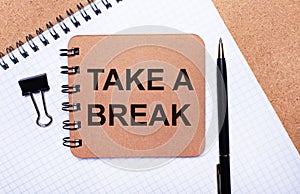 On a wooden background notepad, black pen, paper clip and brown notepad with the text TAKE A BREAK. Business concept